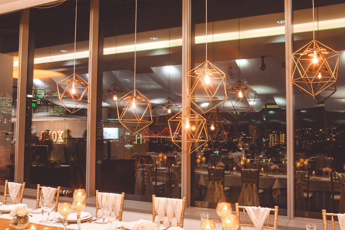 Geometric gold pendant lights above a wedding table at Room Three Sixty with the Story Bridge lit up across the Brisbane skyline at night